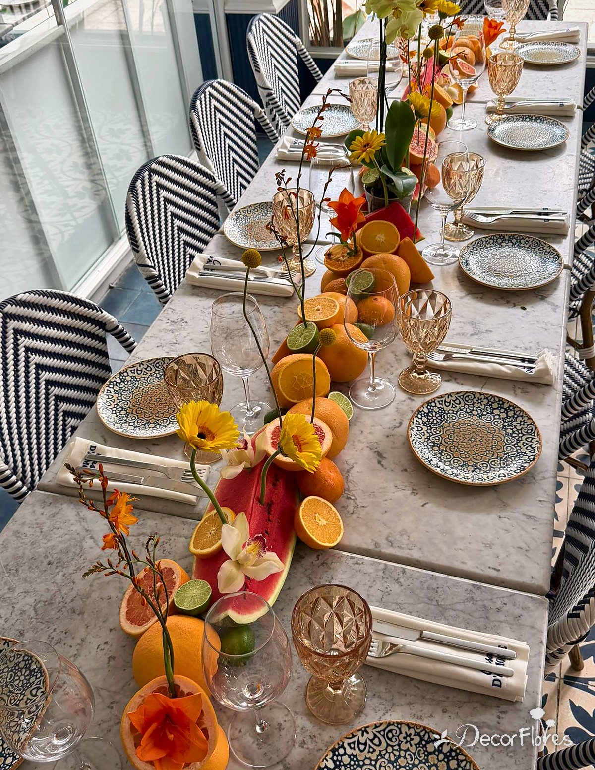 Mesa larga decorada con naranjas, toronjas, limones y patilla partidos, con gerberas, lirios y orquídeas saliendo de la fruta